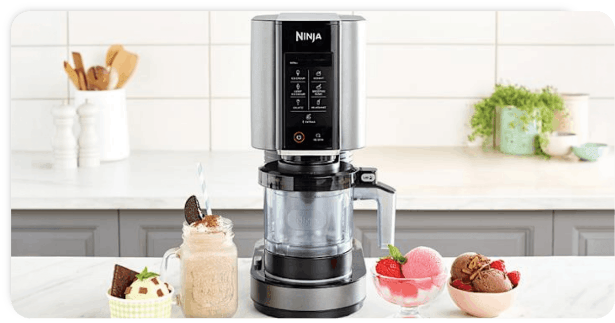 Παγωτομηχανή Ninja με ψηφιακή οθόνη αφής, περιτριγυρισμένη από παγωτά βανίλιας, φράουλας και σοκολάτας καθώς και milkshake σε γυάλινη κούπα, σε λευκή μοντέρνα κουζίνα